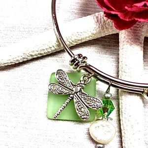 May include: A silver bangle bracelet with a green glass dragonfly charm, a white pearl charm, and a green crystal charm.
