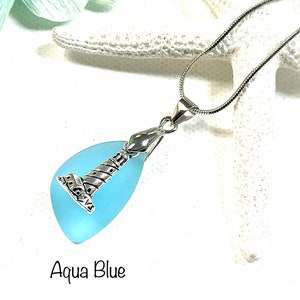 May include: A silver lighthouse charm pendant on a silver chain with a light blue sea glass teardrop. The sea glass is translucent and has a light blue color. The pendant is labeled "Aqua Blue".