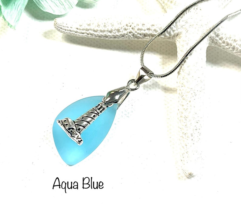 May include: A silver lighthouse charm pendant on a silver chain with a light blue sea glass teardrop. The sea glass is translucent and has a light blue color. The pendant is labeled "Aqua Blue".