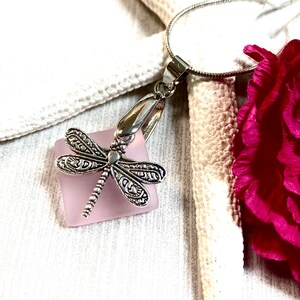 May include: A silver dragonfly charm pendant hangs from a silver chain. The charm is attached to a square piece of light pink glass.