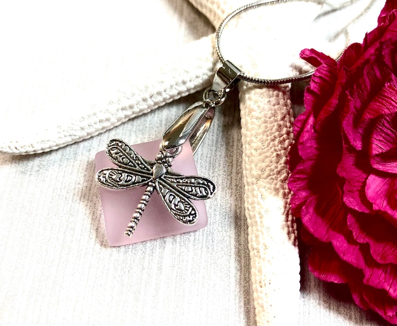 May include: A silver dragonfly charm pendant hangs from a silver chain. The charm is attached to a square piece of light pink glass.