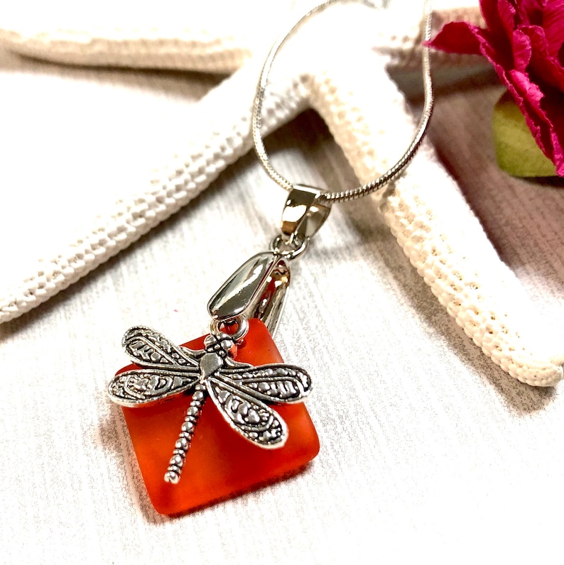 May include: A silver chain necklace with a square piece of orange sea glass and a silver dragonfly charm.