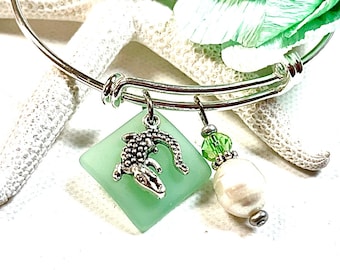 Puede incluir: Un brazalete expandible de plata con un dije de vidrio verde con un delfín plateado, una cuenta de cristal verde y una perla blanca.