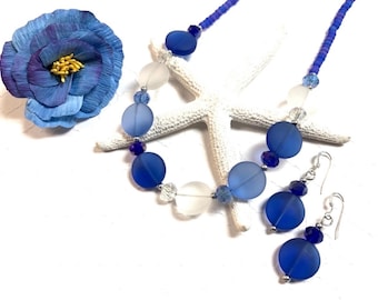 May include: A blue and white glass bead necklace and matching earrings. The necklace features large, round, frosted blue beads, smaller clear beads, and tiny blue beads. The earrings are dangle earrings with a large, round, frosted blue bead, a smaller clear bead, and a tiny blue bead.