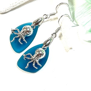 Könnte beinhalten: Ein Paar blaue Glasearrings mit silbernen Kraken-Anhängern. Die Ohrringe sind tropfenförmig und haben eine matte Oberfläche.