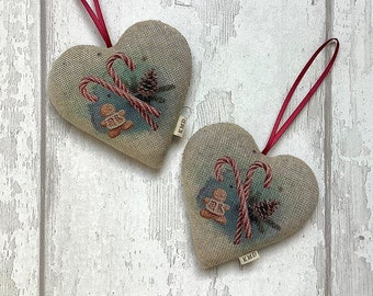 Puede incluir: Dos adornos beige en forma de corazón con cintas rojas. Cada adorno presenta un diseño impreso de bastones de caramelo, un hombre de jengibre y una piña. Los adornos tienen una pequeña etiqueta marrón con el texto "KMD" impreso en ellos.