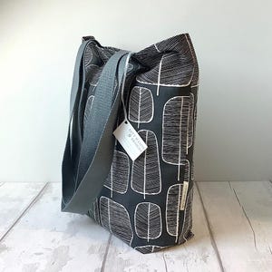 May include: A fabric tote bag featuring a pattern of white leaves on a dark grey background. It has grey straps and a tag reading "Katy Mason Designs". The bag is designed for carrying items and has a simple, modern aesthetic.