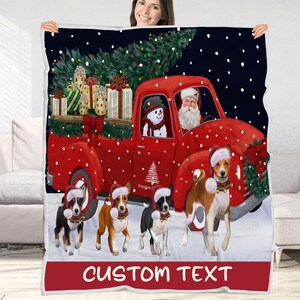 May include: A fleece blanket featuring a red truck carrying a Christmas tree and gifts, with Santa Claus and a snowman in the cab. Four dogs wearing Santa hats stand in front of the truck against a snowy background. The text CUSTOM TEXT is at the bottom.