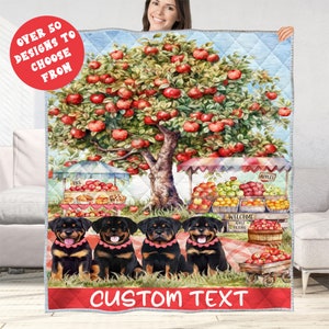 May include: A quilted blanket featuring a vibrant apple orchard scene with a large tree laden with red apples. Below, four Rottweiler dogs sit in a row. The blanket includes the text "CUSTOM TEXT" and "OVER 50 DESIGNS TO CHOOSE FROM".