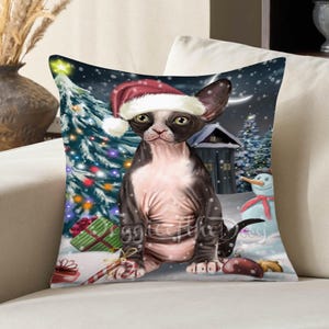 May include: A square throw pillow featuring a graphic illustration of a hairless cat wearing a red and white Santa hat. The scene includes a decorated Christmas tree, a snowman, wrapped gifts, and a candy cane against a snowy night background.