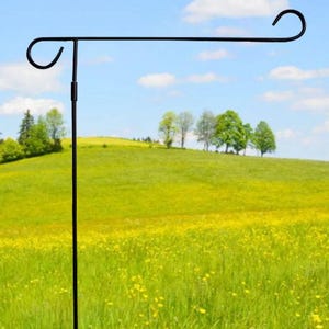 May include: Black metal garden flag stand with a curved arm and a hook at each end. The stand is shown in a field of green grass with yellow wildflowers.
