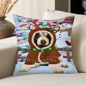 May include: A square throw pillow featuring a graphic illustration of a dog wearing a brown hooded coat with reindeer antlers, a green collar with bells, and buttons. The background shows a snowman, a Christmas tree, and a red cardinal bird.