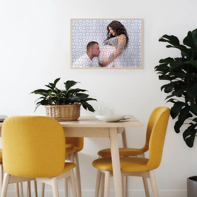 Puede incluir: Un rompecabezas enmarcado con una fotograf&iacute;a de una persona embarazada y otra persona. El rompecabezas se exhibe en una pared blanca sobre una mesa de madera con sillas amarillas y una planta en maceta.