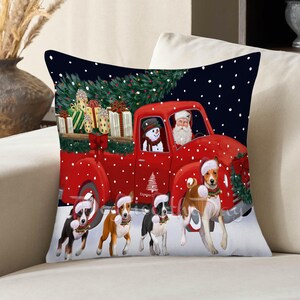 May include: A square throw pillow featuring a festive illustration of a red pickup truck carrying wrapped gifts and a Christmas tree. Santa Claus and a snowman sit in the cab. Four dogs wearing Santa hats stand in the snow in front of the truck.