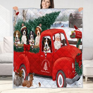 May include: A quilted blanket featuring a red truck driven by Santa Claus, with several dogs in the truck bed. The blanket has a winter scene with a barn, trees, and the text "Happy Holidays".