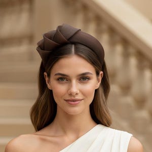 May include: A woman wearing a dark brown headband with a rose-like design. The headband sits atop the head, with the rose detail centered. The woman is wearing a white dress. The background is a blurred outdoor setting.