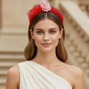 May include: A bright red headband featuring a sheer tulle overlay and a single pale pink rose accent positioned on the top. The headband has a smooth, rounded structure and is designed to sit securely across the crown of the head.