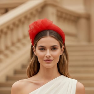 Puede incluir: Una diadema roja con un diseño floral en capas. La persona lleva un vestido blanco de un solo hombro. Al fondo, una escalera.