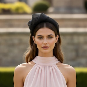 Op de afbeelding: Een zwarte geweven hoofdband met een gestructureerd ontwerp. De hoofdband wordt gedragen met een lichtroze haltertopjurk. De achtergrond toont een vage buitenomgeving met groen en een gebouw.