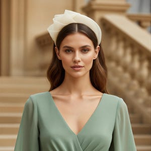 May include: A woman wearing a cream-colored headpiece with a rose-like design. The headpiece sits atop the woman's head, and she is wearing a sage green dress with a V-neckline and long sleeves. The background is a blurred outdoor setting.