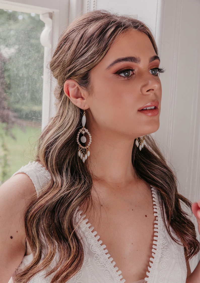 May include: Gold-coloured chandelier earrings with white and pearl details. The earrings have a circular design with dangling teardrop beads. The model is wearing a white dress with a lace trim.