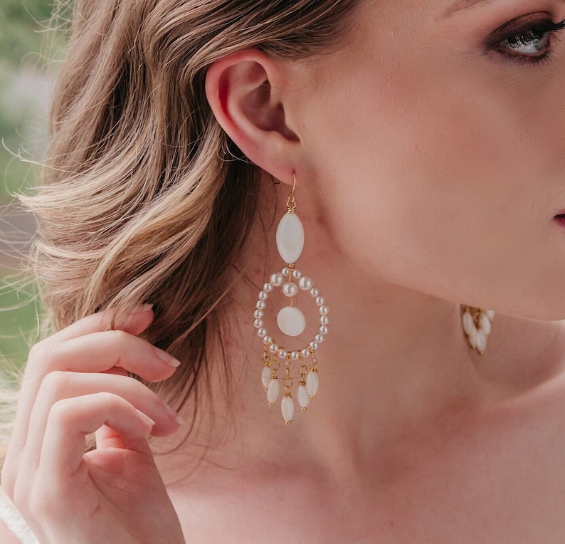 May include: Elegant gold-tone earrings with white oval and teardrop-shaped accents. The earrings feature a circular design with small pearls and dangling teardrops, perfect for special occasions.