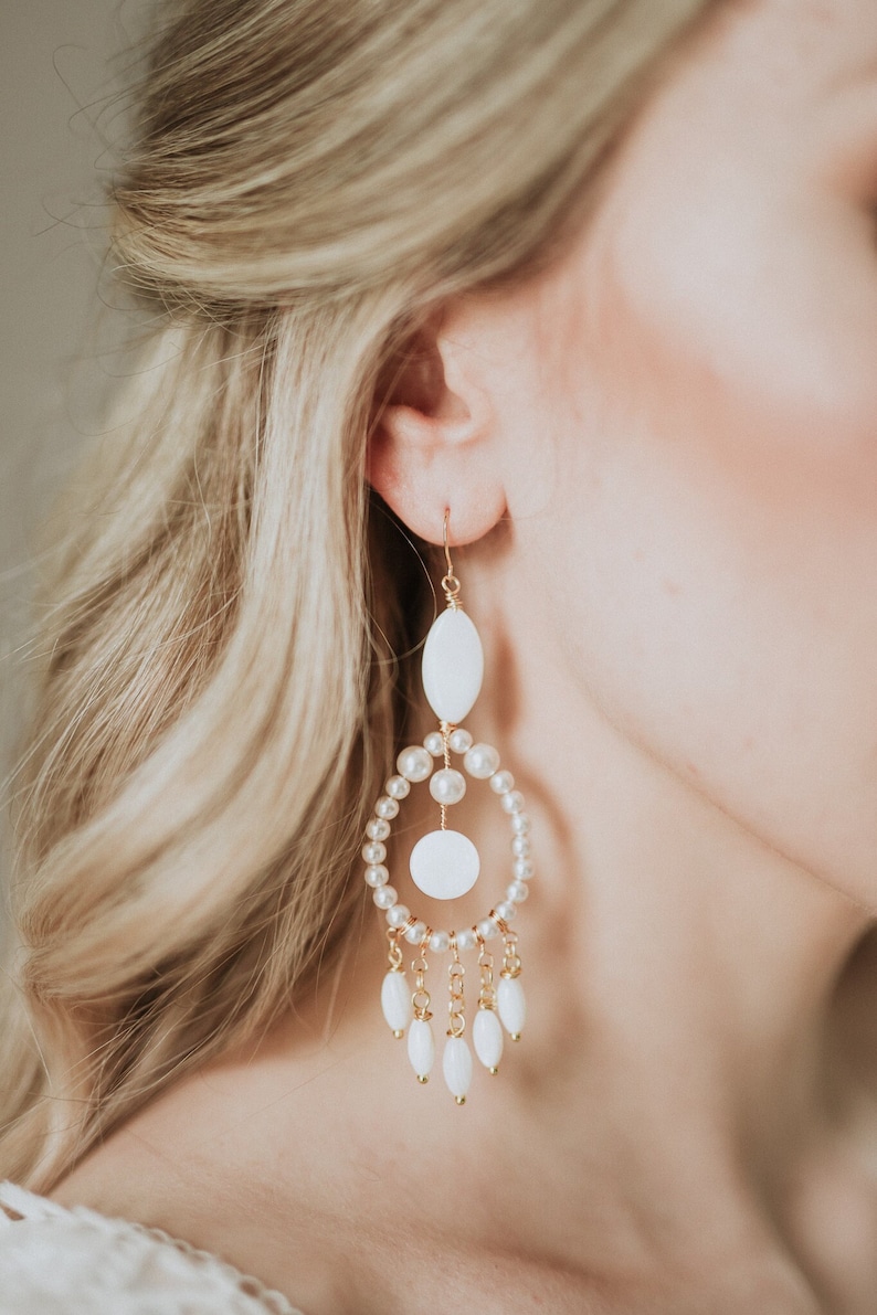 May include: Elegant gold-tone earrings with white oval and round accents, and dangling white teardrop-shaped beads. The earrings feature a pearl-lined hoop design, creating a sophisticated and stylish accessory. Perfect for special occasions.