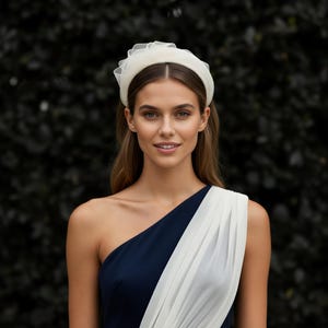 May include: A woman wearing a white headband with a sheer, ruffled trim. She is wearing a one-shoulder dress with a navy blue bodice and a white draped overlay. The background is a dark green hedge.