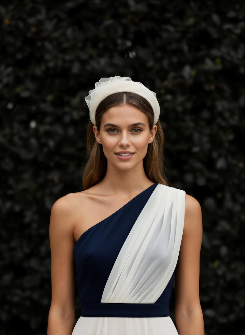 May include: A woman wearing a white headband with a sheer, ruffled trim. She is wearing a one-shoulder dress with a navy blue bodice and a white draped overlay. The background is a dark green hedge.