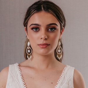 May include: A woman wearing gold-toned chandelier earrings with white and pearl accents. The earrings have a teardrop shape and dangle from the ear. The woman is wearing a white, sleeveless top with a detailed, embroidered design.