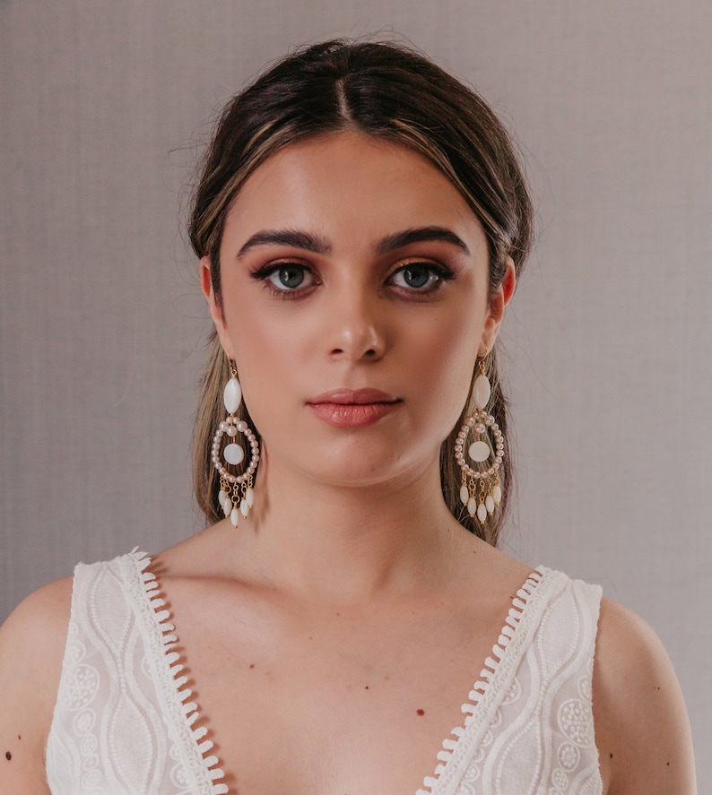 May include: A woman wearing gold-toned chandelier earrings with white and pearl accents. The earrings have a teardrop shape and dangle from the ear. The woman is wearing a white, sleeveless top with a detailed, embroidered design.
