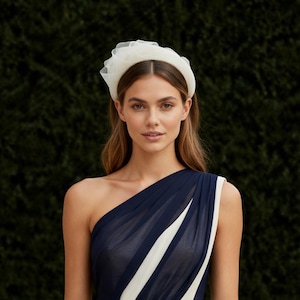 May include: A woman wearing a white tulle headband and a navy blue one-shoulder dress with a white stripe. The headband is a wide band with a ruffled tulle accent. The dress has a flowing design.