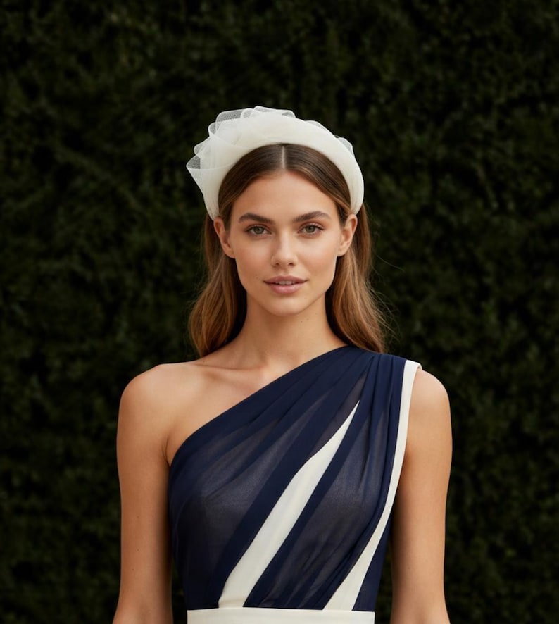 May include: A woman wearing a white tulle headband and a navy blue one-shoulder dress with a white stripe. The headband is a wide band with a ruffled tulle accent. The dress has a flowing design.