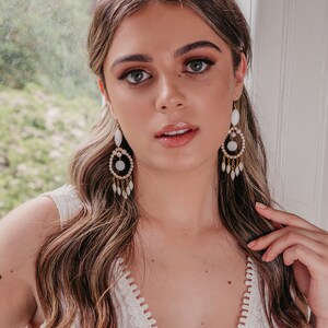 May include: A woman wearing white and gold chandelier earrings. The earrings have a circular design with white stones and dangling teardrop-shaped accents. The woman is wearing a white dress and has long, wavy brown hair.