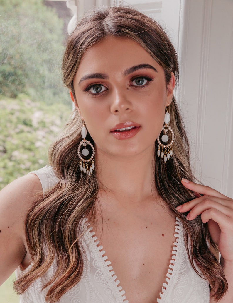 May include: A woman wearing white and gold chandelier earrings. The earrings have a circular design with white stones and dangling teardrop-shaped accents. The woman is wearing a white dress and has long, wavy brown hair.