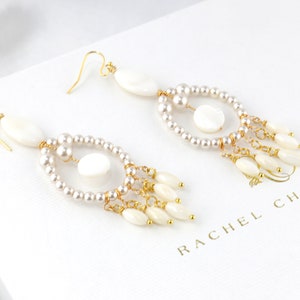May include: Elegant dangle earrings featuring white oval and round shell accents, complemented by small pearls and gold-tone metal detailing. The earrings have a delicate, layered design with a cascading effect. The brand name "RACHEL CHAPRUN" is visible.