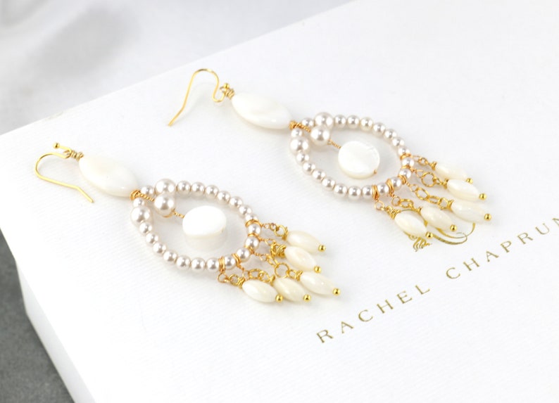 May include: Elegant dangle earrings featuring white oval and round shell accents, complemented by small pearls and gold-tone metal detailing. The earrings have a delicate, layered design with a cascading effect. The brand name "RACHEL CHAPRUN" is visible.