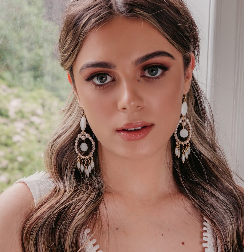 May include: Elegant chandelier earrings with a pearl and gold design. The earrings feature a large oval-shaped pearl at the top, a circular design with pearls, and dangling pearl accents. The earrings are a classic accessory.