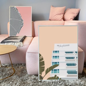 Pu&ograve; includere: Una stampa incorniciata di un edificio bianco con il testo "The Carlyle" sul tetto. L'edificio &egrave; su uno sfondo pesca con palme in primo piano.