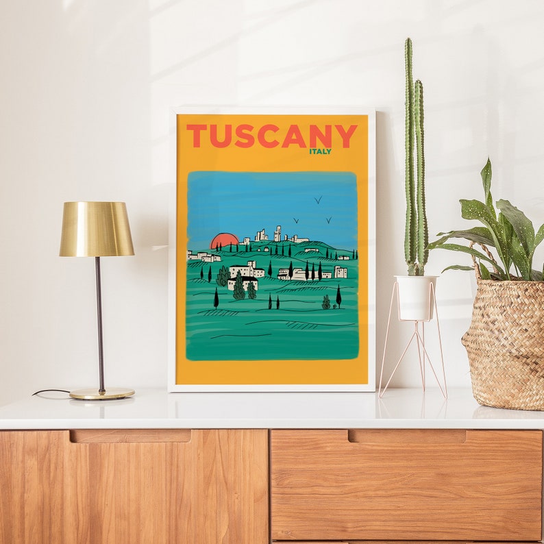 K&ouml;nnte beinhalten: Ein wei&szlig; gerahmtes Poster mit dem Text "Tuscany, Italy" in roten und schwarzen Buchstaben. Das Poster zeigt eine stilisierte Illustration einer toskanischen Landschaft mit einer Stadt in der Ferne, gr&uuml;nen H&uuml;geln und einer roten Sonne, die hinter den H&uuml;geln untergeht.