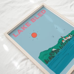 May include: A framed illustration of Lake Bled in Slovenia. The illustration features a blue lake, green mountains, a church on an island, and a red sun in a blue sky. The text "LAKE BLED" is written in pink letters at the top of the illustration.