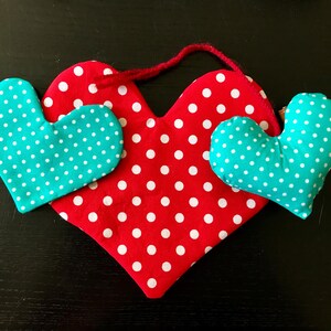 May include: A handmade heart-shaped decoration with a red polka-dot pattern and two smaller teal hearts with white polka dots. A red string is attached for hanging. The hearts are made of fabric and are likely stuffed.