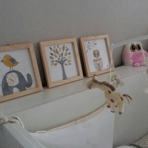 May include: Three framed nursery art prints with animal illustrations: an elephant with a bird, a tree, and an owl. A baby mobile with plush animal figures hangs nearby. A pink owl plush toy is also visible.