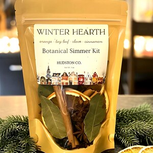 May include: A tan stand-up pouch labeled Winter Hearth Botanical Simmer Kit by Hudston Co. The label lists orange, bay leaf, clove, and cinnamon. The clear window reveals dried orange slices, bay leaves, cinnamon sticks, and star anise inside.