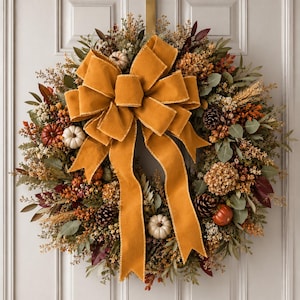 May include: A decorative wreath featuring a large mustard yellow velvet bow, small white pumpkins, brown pinecones, dried foliage, and orange berries. The arrangement is circular, composed of various autumn-toned dried plants and greenery.