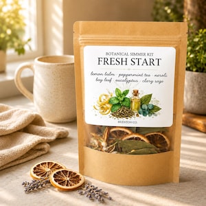 May include: A brown paper bag with a clear window, labeled "FRESH START" with botanical ingredients. The bag contains dried orange slices, bay leaves, and other herbs. A white mug and a beige cloth are in the background. Dried lavender and orange slices are in the foreground.