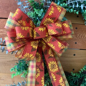 May include: A decorative wreath with a large bow featuring sunflowers. The bow has a burnt orange background with yellow sunflowers and brown leaves. The edges of the bow are a plaid pattern of yellow, green, and orange. The wreath has green foliage.