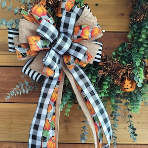 May include: A fall-themed wreath with a large bow featuring pumpkin and buffalo check patterns. The bow is made of burlap and black and white striped ribbon. The wreath includes green foliage and a small jack-o'-lantern decoration.
