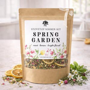 May include: A brown paper bag labeled "Stovetop Simmer Kit Spring Garden" with a clear window showing dried herbs, lemon slices, and flowers. The bag is tied with a white ribbon. Dried lemon slices and fresh herbs are arranged around the bag.
