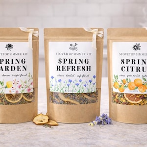 May include: Three brown paper pouches labeled "Stovetop Simmer Kit" with "Spring Garden", "Spring Refresh", and "Spring Citrus" text. Each pouch has a clear window showing dried herbs and fruit. The "Spring Garden" pouch has floral accents.